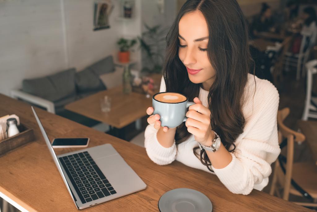 молодая привлекательная женщина-фрилансер пьет кофе за столом с ноутбуком и смартфоном в кафе
  - Фото, изображение