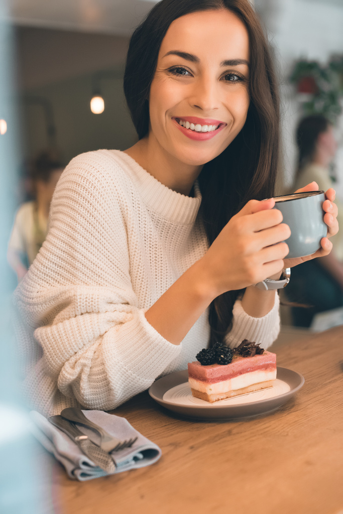 s úsměvem mladá žena s šálek kávy sedí u stolu s tvarohový koláč v kavárně - Fotografie, Obrázek