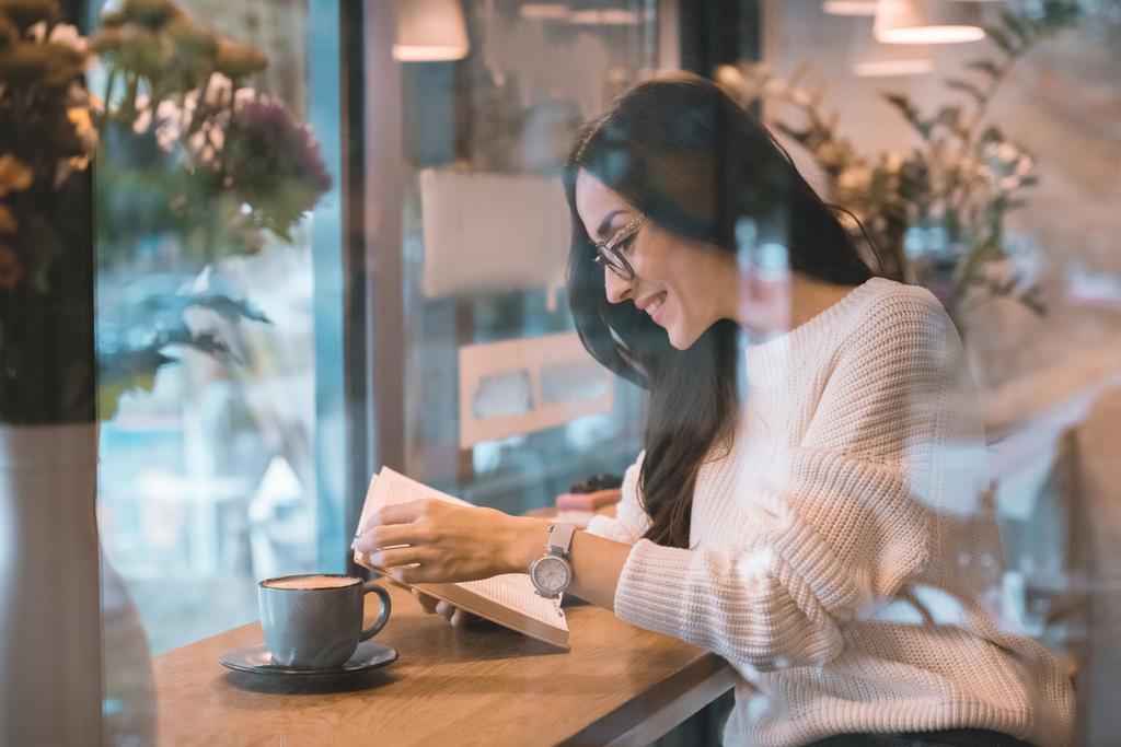 χαμογελαστή γυναίκα ανάγνωση βιβλίων στο τραπέζι με φλιτζάνι καφέ στην καφετέρια - Φωτογραφία, εικόνα
