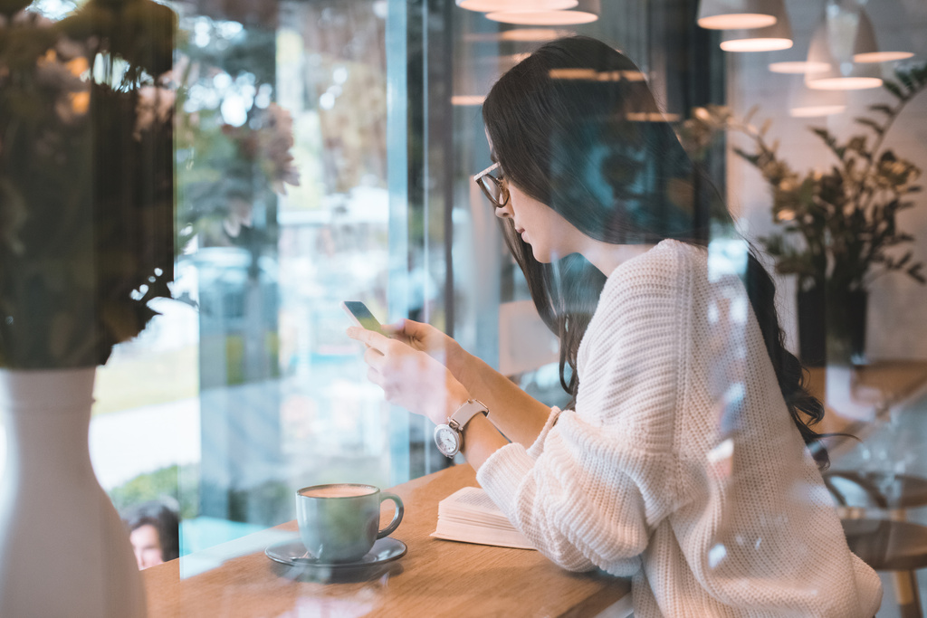 Widok z boku z młodą kobietę w okularach za pomocą smartphone przy stole z filiżanki kawy w kawiarni - Zdjęcie, obraz