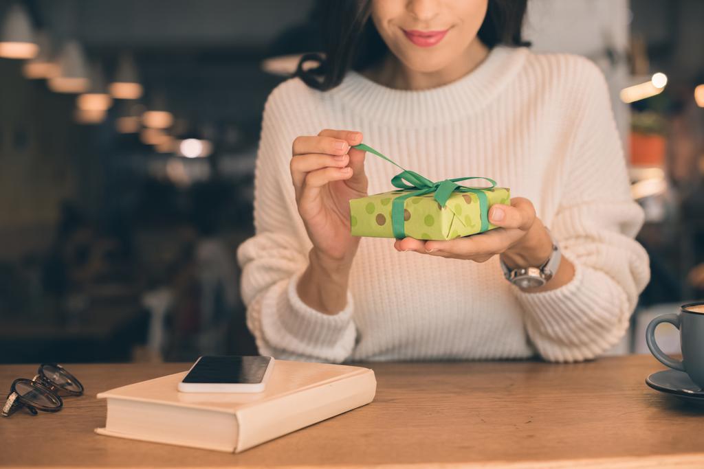 vista parcial de la mujer joven desatando caja de regalo en la mesa con teléfono inteligente y taza de café en la cafetería
 - Foto, Imagen