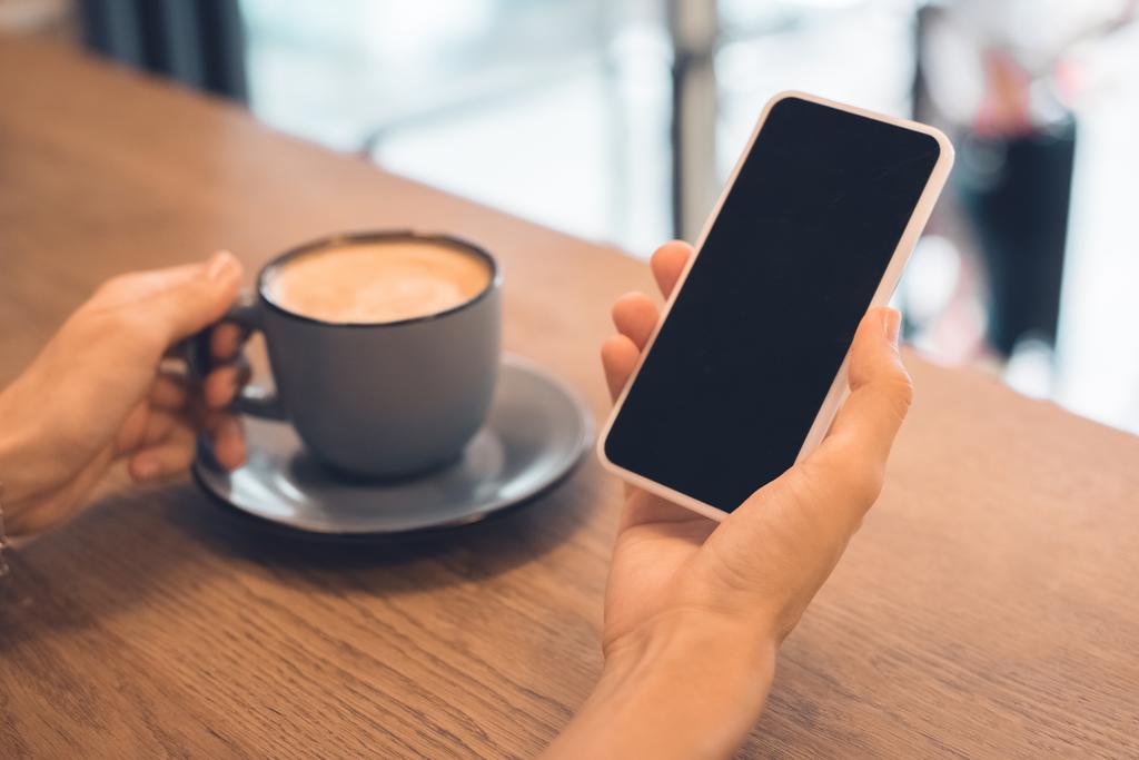 обрезанный образ женщины с чашкой кофе с помощью смартфона с пустым экраном за столом в кафе
 - Фото, изображение
