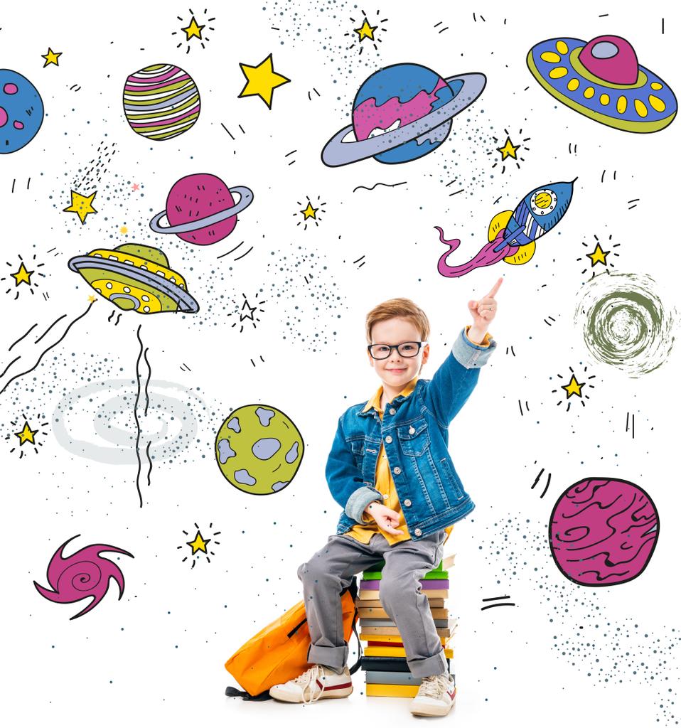 smiling schoolboy pointing up while sitting on pile of books with backpack, isolated on white with fantasy universe, planets and spaceships - Photo, Image
