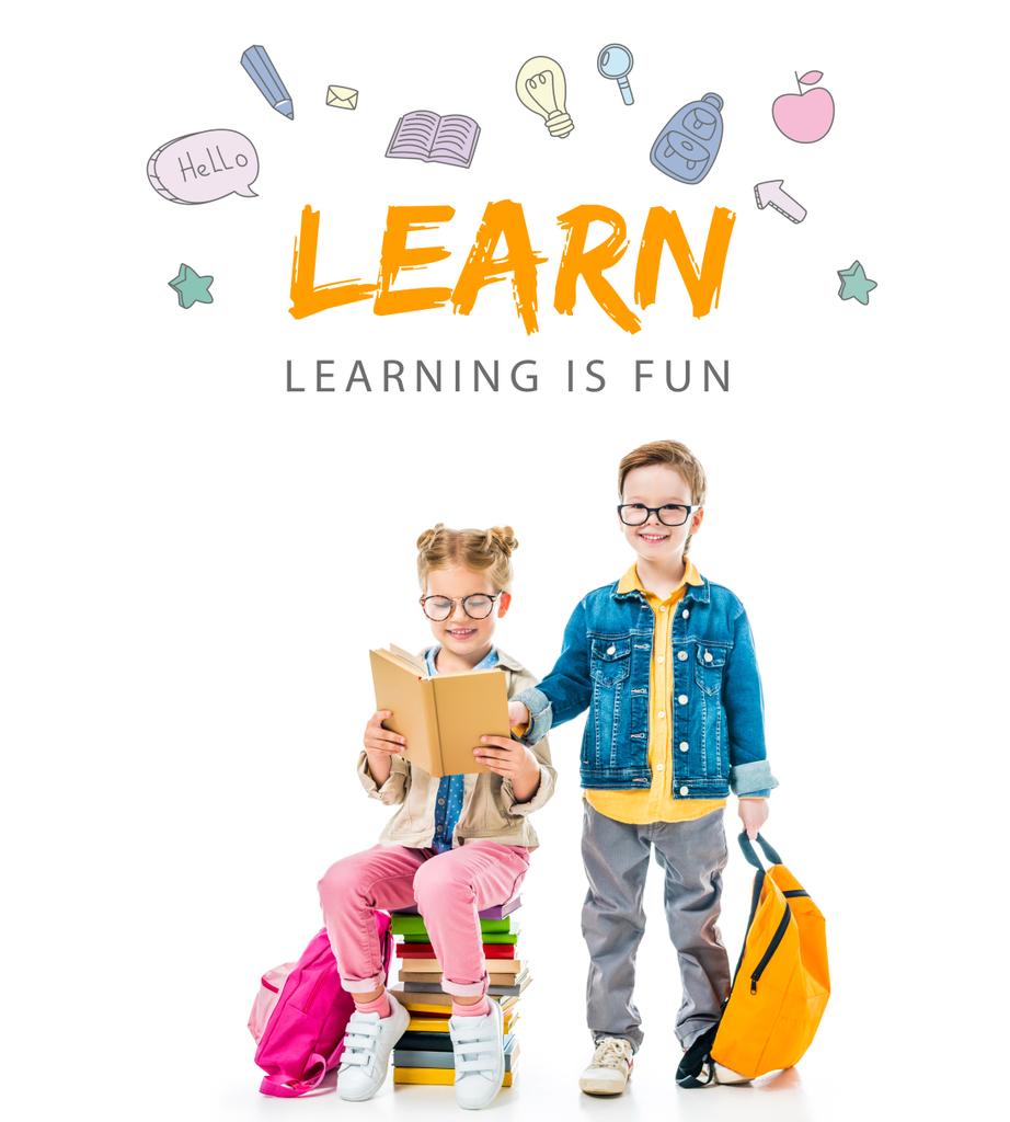 schoolkinderen in brillen studeren en zittend op boeken met rugzakken, geïsoleerd op wit met "leren - leren is leuk" belettering - Foto, afbeelding