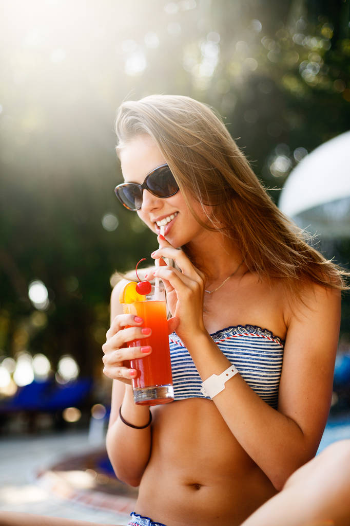 Ritratto di giovane donna con bicchiere da cocktail che si rilassa al sole tropicale vicino alla piscina su una sedia a sdraio con le palme dietro. Concetto di vacanza
 - Foto, immagini