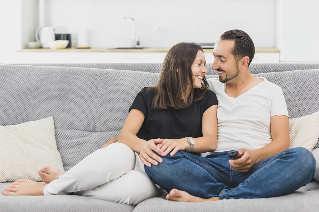 Die glückliche Familie schaut fern, während sie auf dem Sofa sitzt. Paar in lässiger Kleidung auf einem modernen grauen Sofa. Liebendes Paar schaut einander an - Foto, Bild