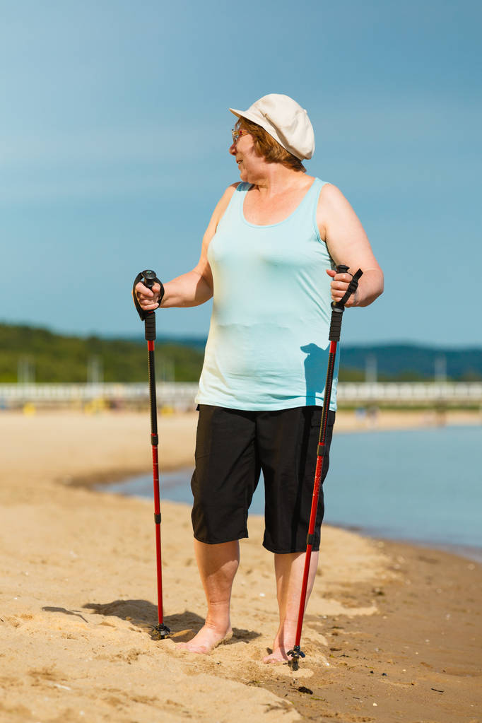 Mulher sênior praticando caminhada nórdica na costa do mar, idosa ativa desfrutando de dia ensolarado de verão. Estilo de vida saudável na velhice
. - Foto, Imagem