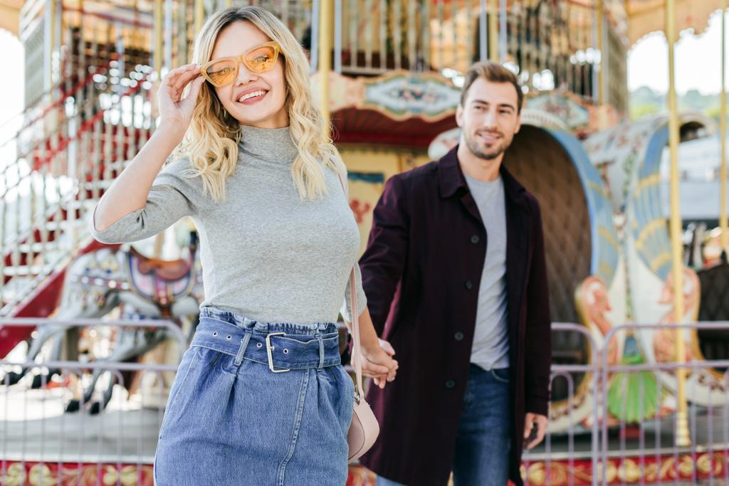 sourire couple affectueux en tenue d'automne tenant la main près du carrousel dans le parc d'attractions, petite amie regardant la caméra
 - Photo, image