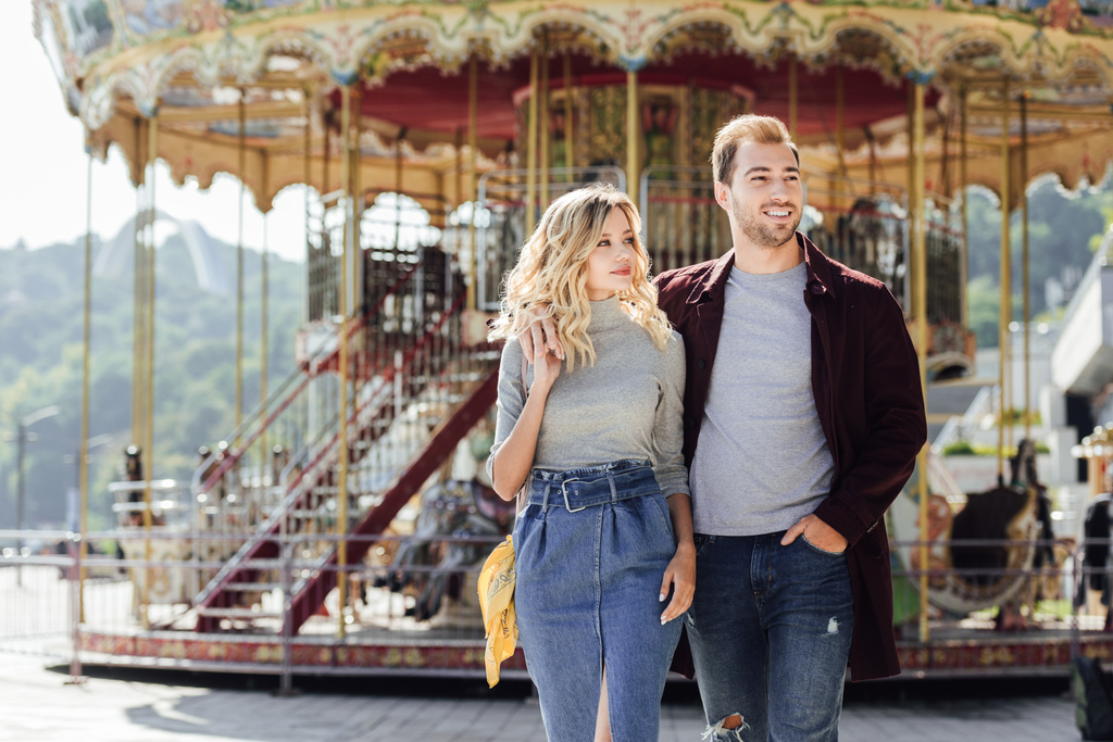 heterosexual couple in autumn outfit walking and cuddling near carousel in amusement park - Photo, Image