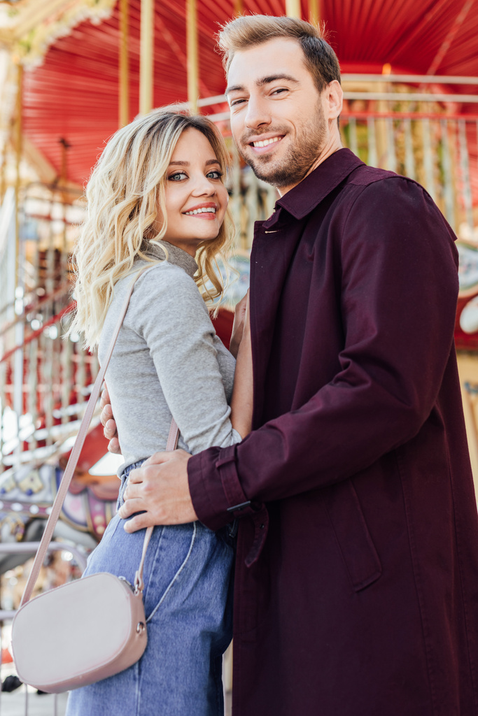 sorrindo casal elegante abraçando perto de carrossel no parque de diversões e olhando para a câmera
 - Foto, Imagem