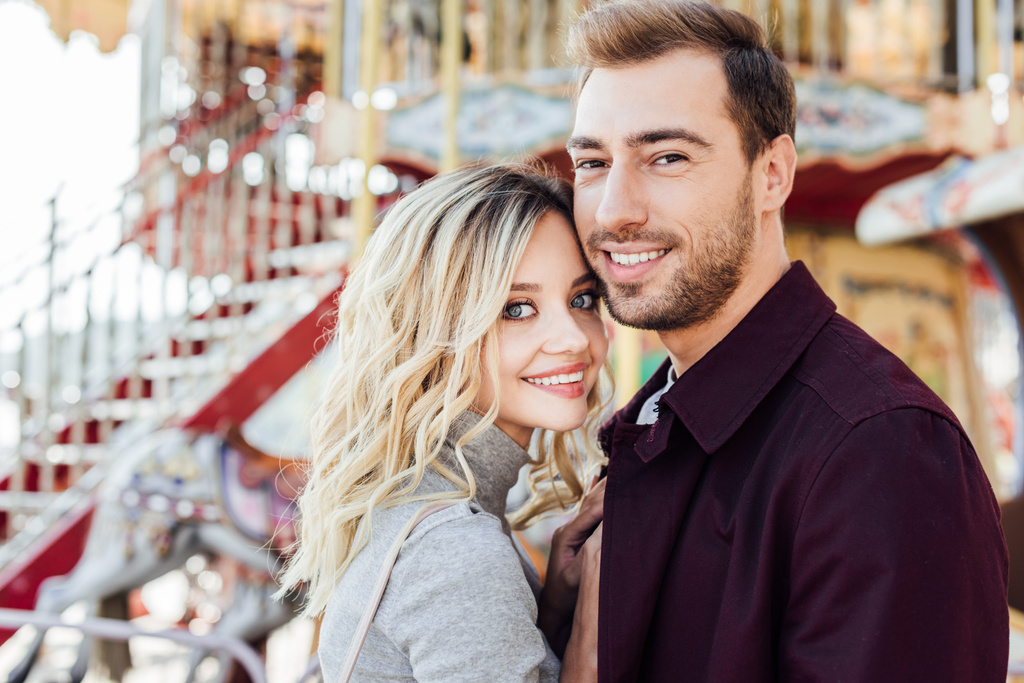 retrato de pareja cariñosa sonriente en traje de otoño mirando la cámara cerca del carrusel en el parque de atracciones
 - Foto, Imagen