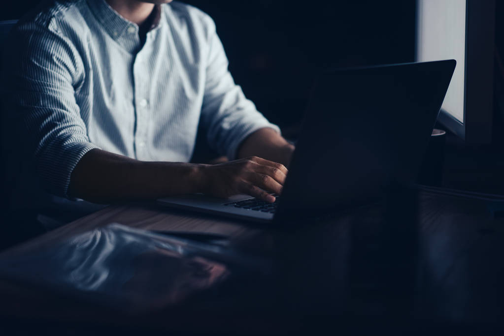 Primer plano de un hombre de negocios sentado en su escritorio en una oficina oscura tarde en la noche trabajando en línea con un ordenador portátil
 - Foto, Imagen