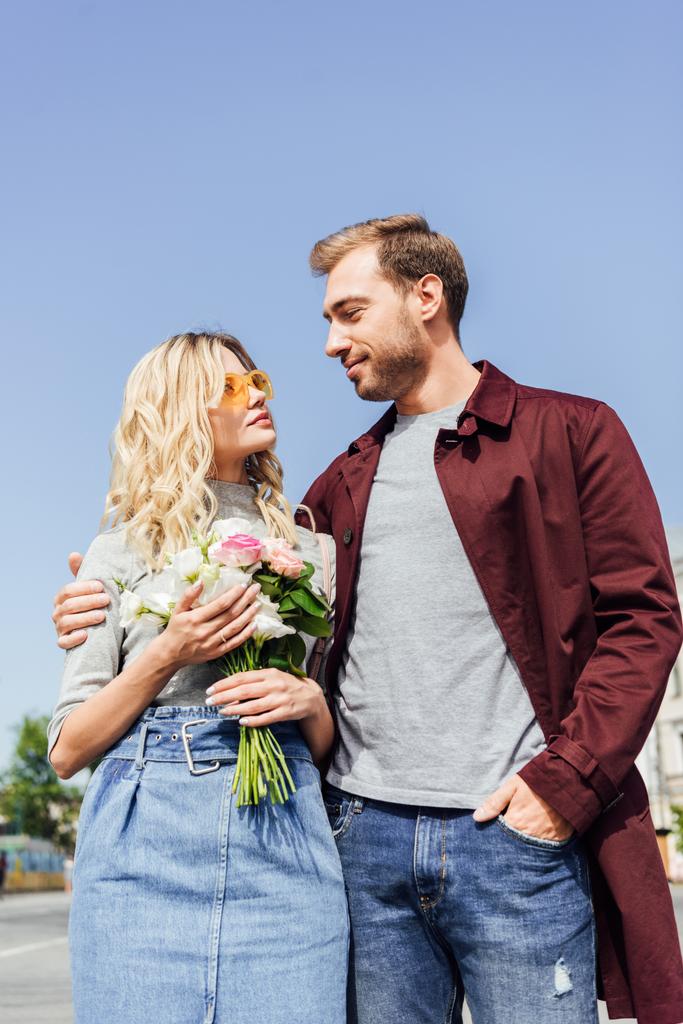 nízký úhel pohledu páru v podzimní oblečení na sebe dívali na ulici, Přítelkyně drží kytici růží - Fotografie, Obrázek