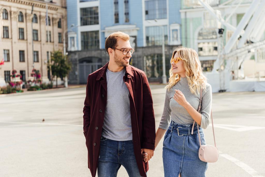 smiling couple in autumn outfit holding hands and looking at each other in city - Photo, Image