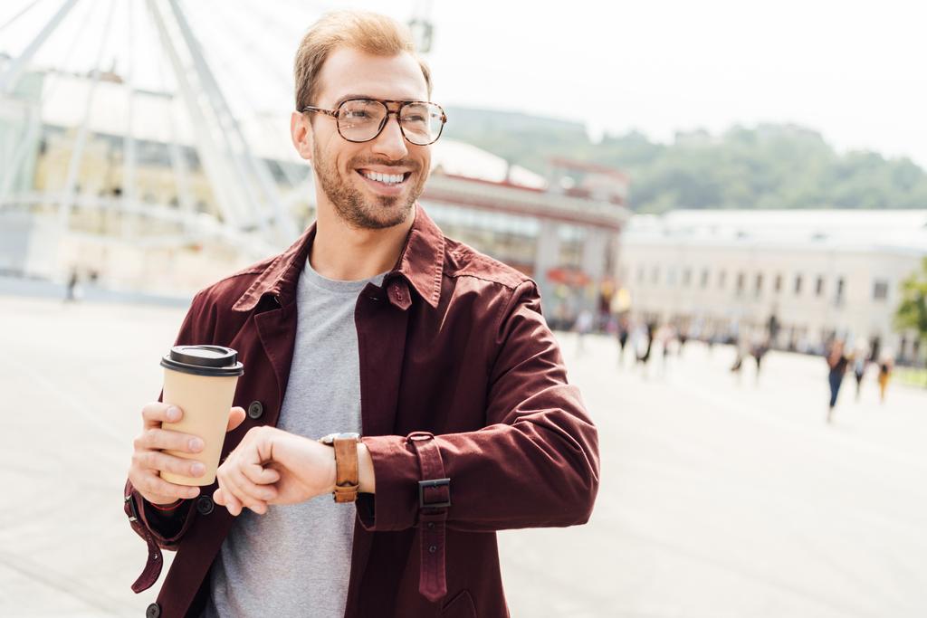 χαμογελώντας όμορφος άνδρας στο φθινόπωρο στολή κρατώντας φλιτζάνι καφέ μίας χρήσης και τον έλεγχο του χρόνου στην πόλη - Φωτογραφία, εικόνα