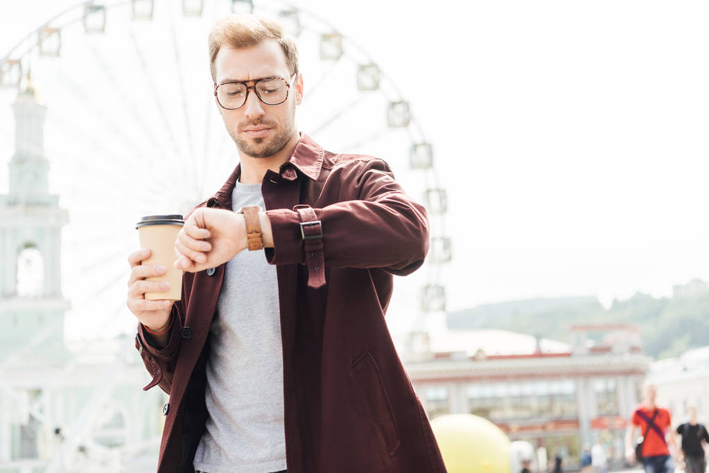 niski kąt widzenia przystojny mężczyzna w stroju jesień gospodarstwa w papierowy kubek kawy i sprawdzanie czasu w pobliżu koło obserwacyjne - Zdjęcie, obraz