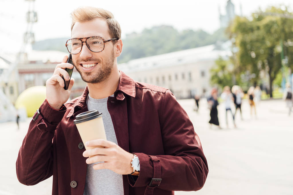 Glimlachende man in herfst-outfit houden wegwerp koffiekopje en praten door smartphone in stad - Foto, afbeelding