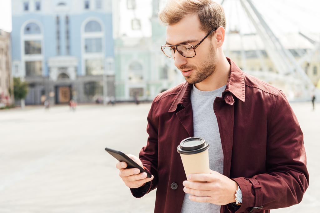 pohledný muž v podzimní oblečení drží kávu jít a použití smartphone v městě - Fotografie, Obrázek