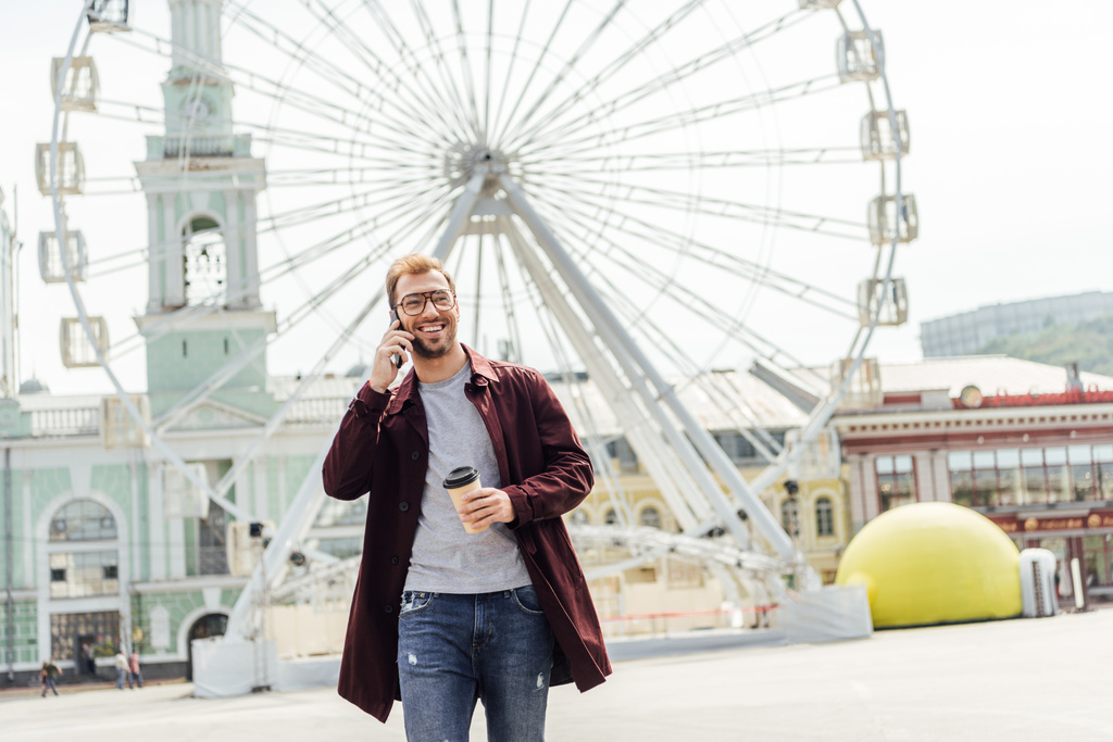 smiling man in autumn outfit holding coffee to go and talking by smartphone near observation wheel - Photo, Image