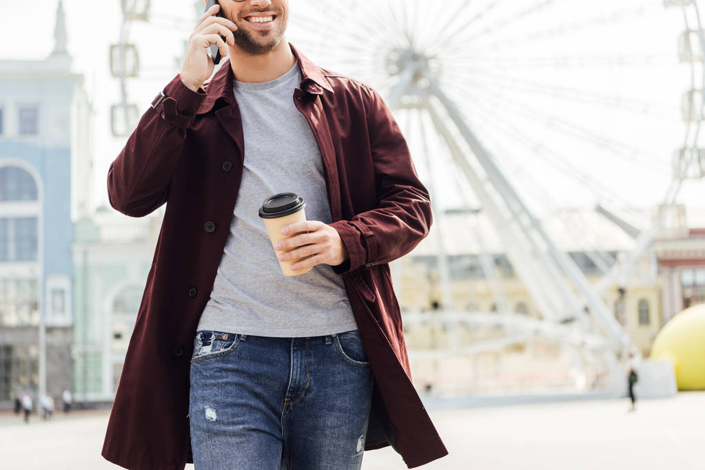 περικομμένη εικόνα του ανθρώπου σε Φθινοπωρινό ντύσιμο κρατώντας καφέ για να πάει και να μιλάμε από το smartphone κοντά ρόδα παρατήρησης - Φωτογραφία, εικόνα