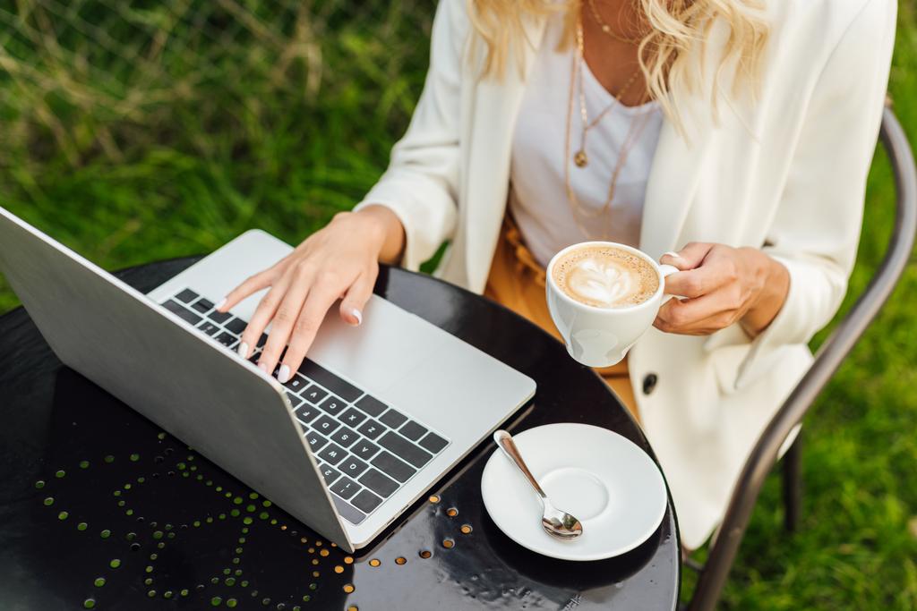 bijgesneden afbeelding van vrouw met laptop en bedrijf kopje koffie aan tafel in de tuin - Foto, afbeelding