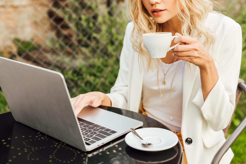 cropped image of woman using laptop and holding cup of coffee at table in garden - Photo, Image
