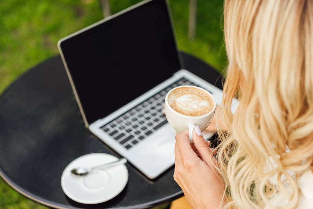 обрезанное изображение женщины с чашкой кофе, ноутбук с пустым экраном на столе в саду
 - Фото, изображение