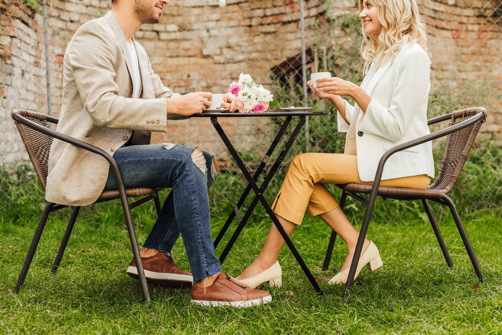 vista laterale della coppia in abito autunnale seduta a tavola in caffè con tazze di caffè e bouquet sul tavolo
 - Foto, immagini