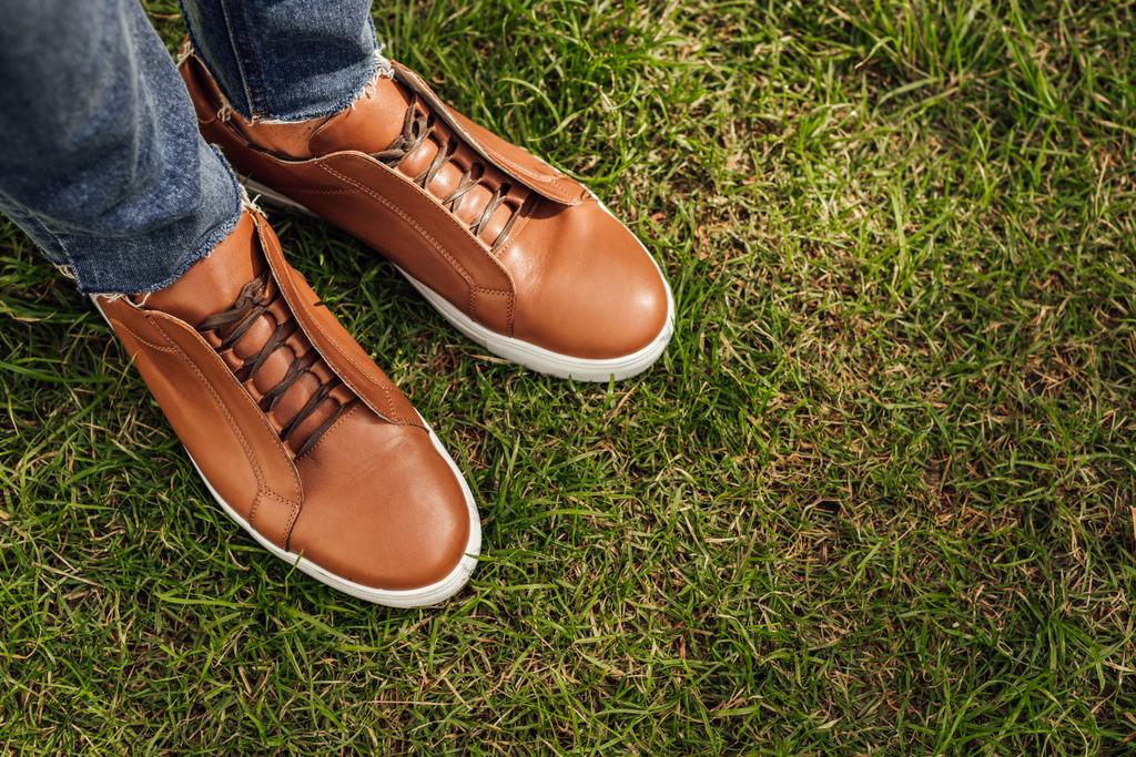 oříznutý obraz člověka v džíny a hnědé boty stojící na zelené trávě v parku - Fotografie, Obrázek