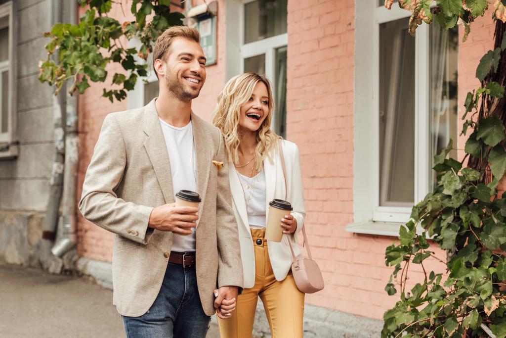 手を繋いでいると使い捨てのコーヒー カップが付いている通りを歩く秋の服装のカップルを笑う - 写真・画像