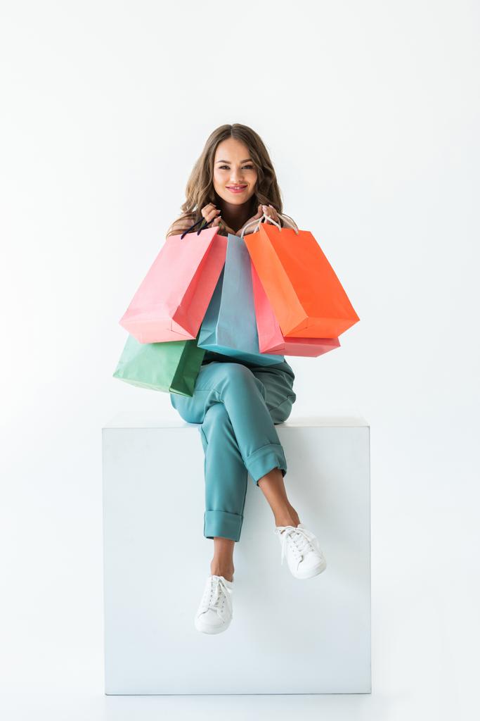 s úsměvem shopaholic sedí na bílé krychle s nákupní tašky, izolované na bílém - Fotografie, Obrázek