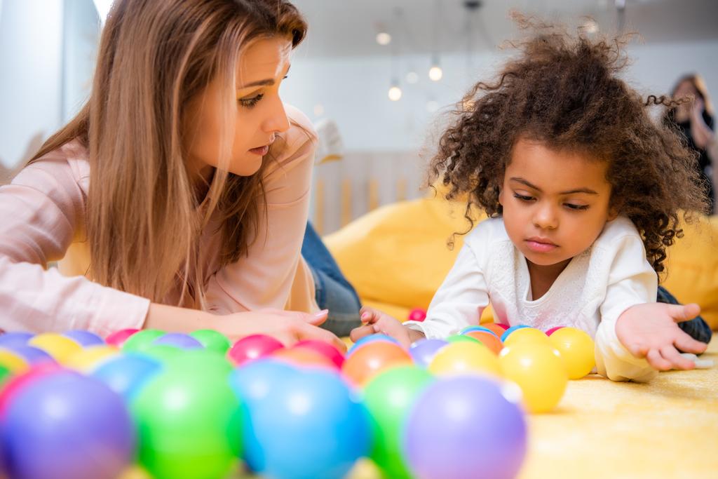 caucasian tutor and adorable african american kid playing with balls in kindergarten - Photo, Image