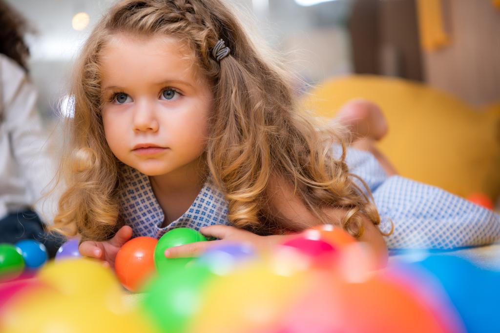 adorável criança deitada no tapete com bolas coloridas no jardim de infância e olhando para longe
 - Foto, Imagem