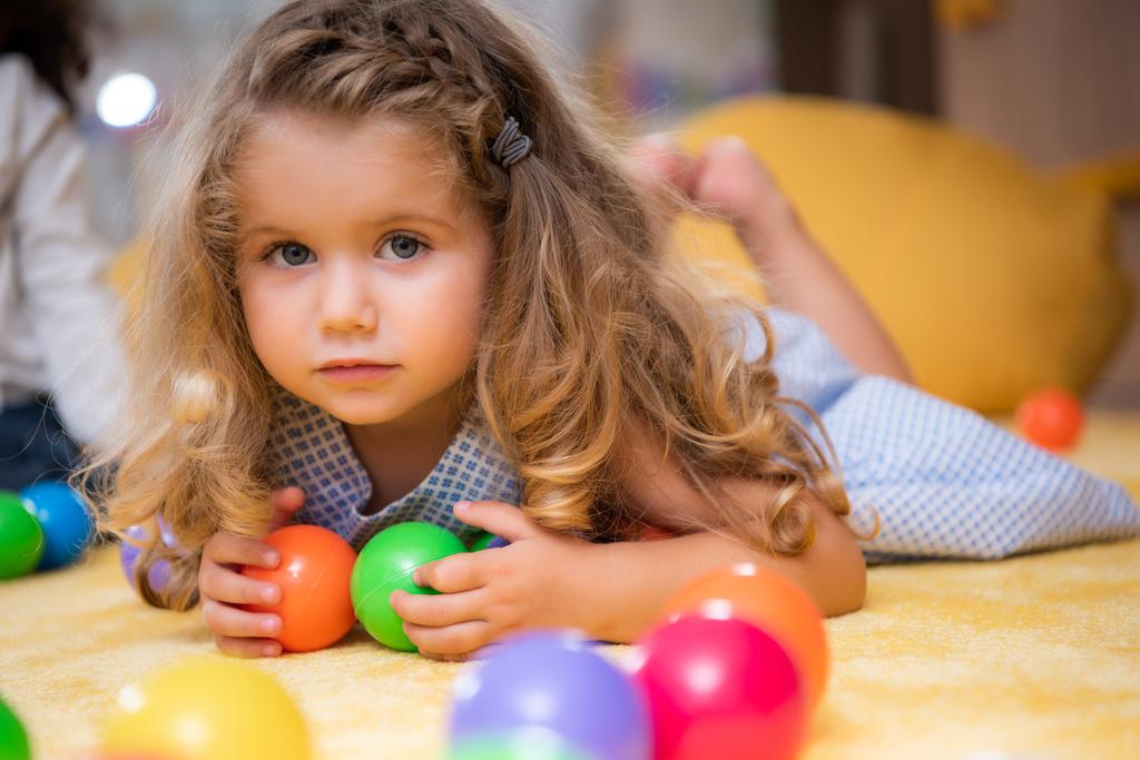 entzückendes Kind liegt mit bunten Kugeln im Kindergarten auf Teppich und blickt in die Kamera - Foto, Bild