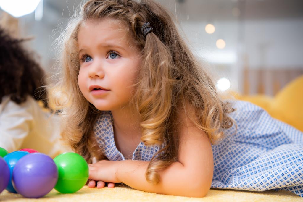 adorable niño acostado en la alfombra con juguetes y mirando hacia otro lado en el jardín de infantes
 - Foto, imagen