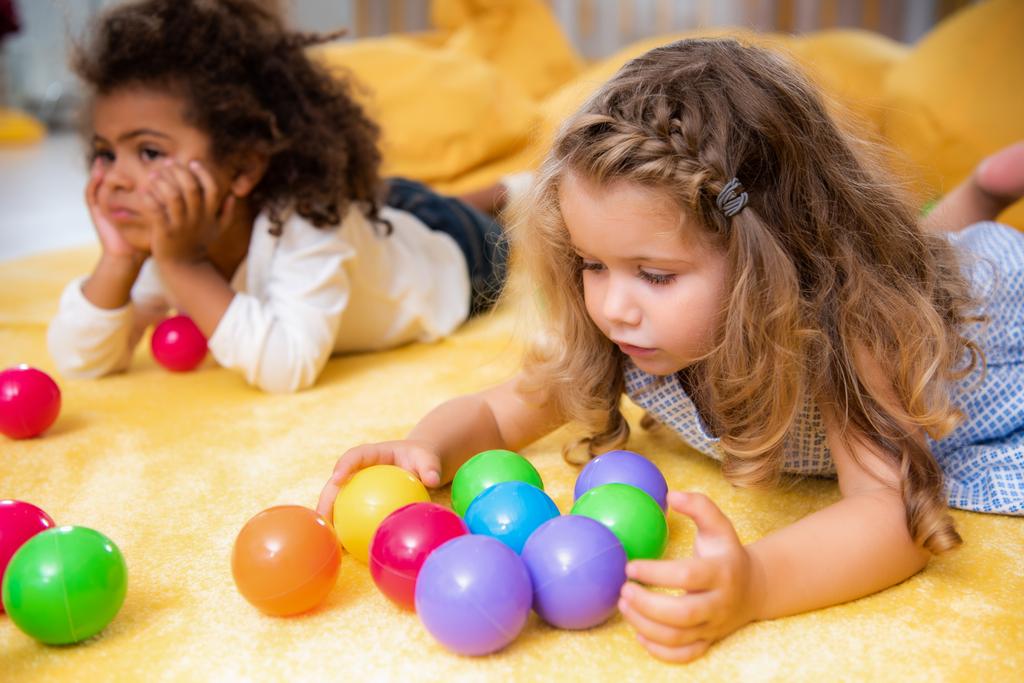 schattig multi-etnisch kinderen spelen op geel tapijt met gekleurde ballen in de kleuterschool - Foto, afbeelding