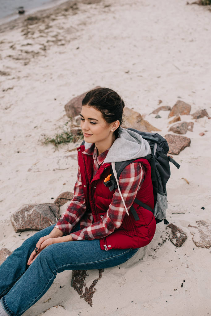 mujer pensativa con mochila descansando sobre rocas en la playa de arena
 - Foto, imagen