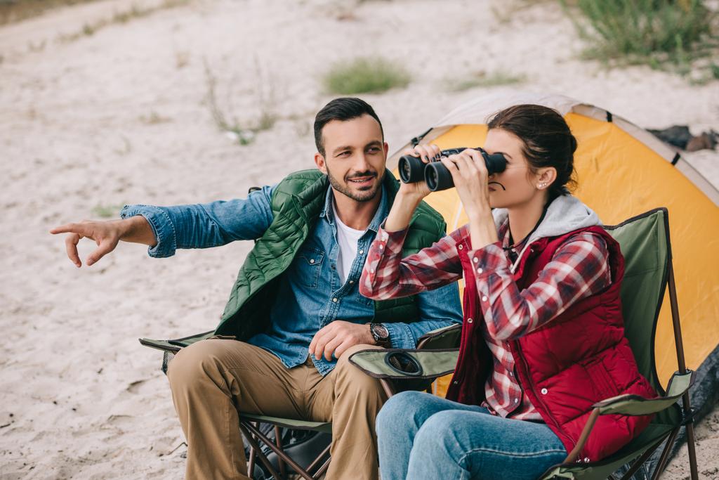 woman looking through binoculars while having camping together with husband on sandy beach - Photo, Image