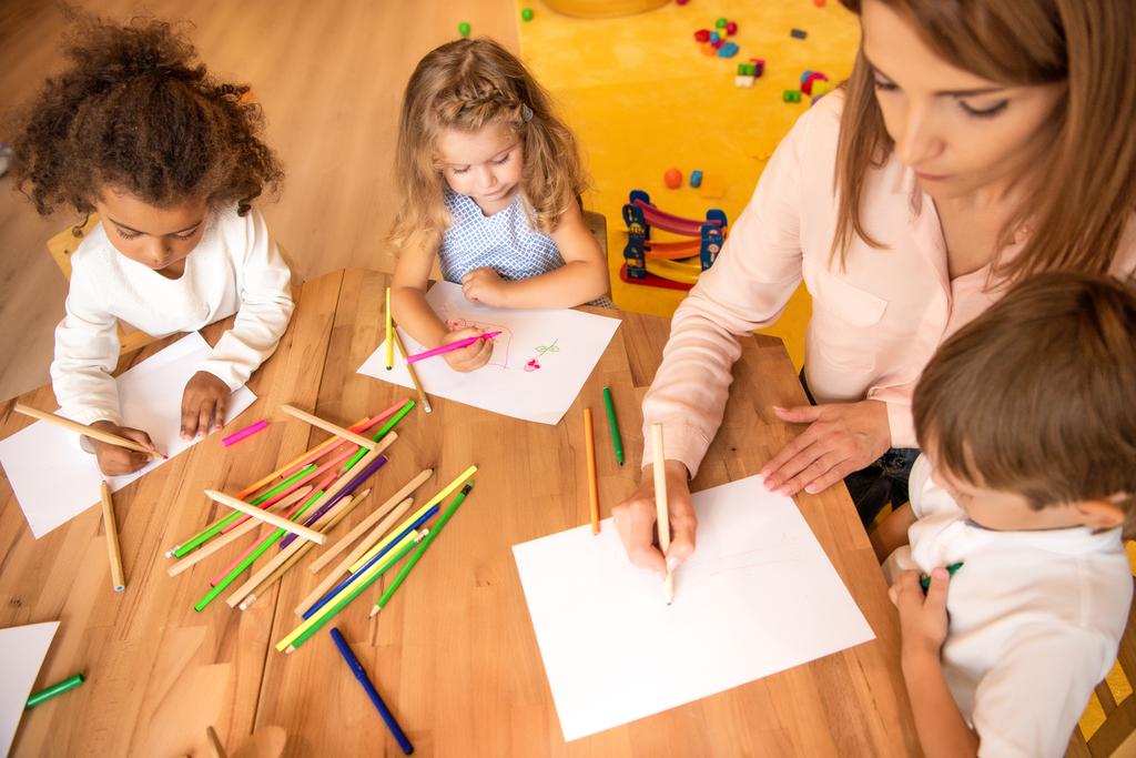высокий угол зрения педагога и мультиэтнических детей рисования в детском саду
 - Фото, изображение