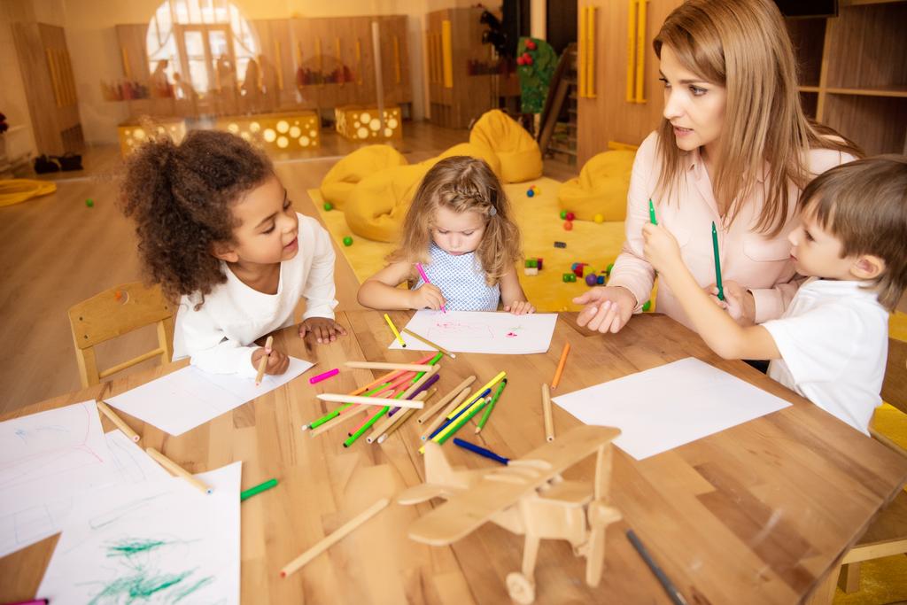 педагог беседует с мультикультурными детьми, рисующими за столом в детском саду
 - Фото, изображение