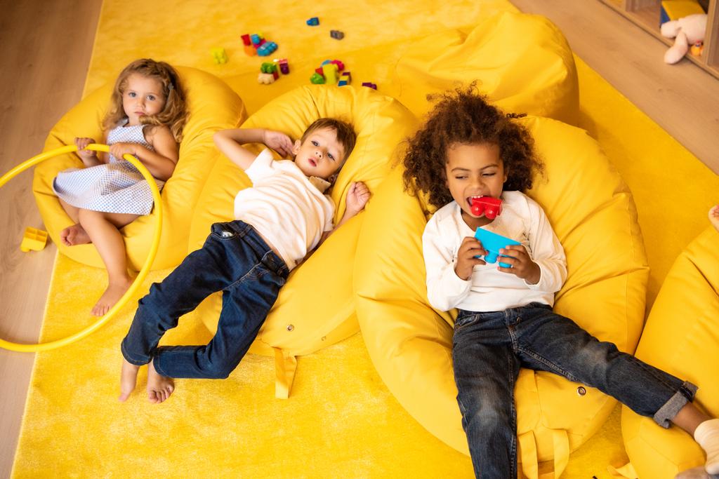pohled z vysoké úhlu mnohonárodnostní děti ležící na židle fazole vak s hračkami v MŠ - Fotografie, Obrázek