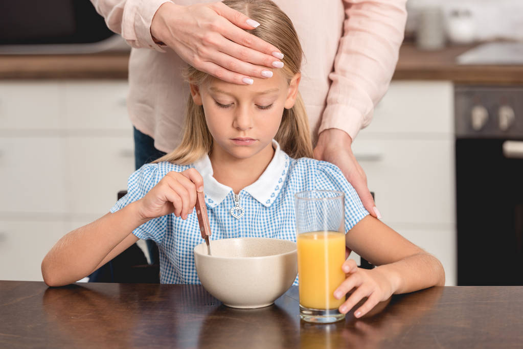 recortado disparo de madre comprobar hijas temperatura con la mano mientras ella desayunando
 - Foto, Imagen