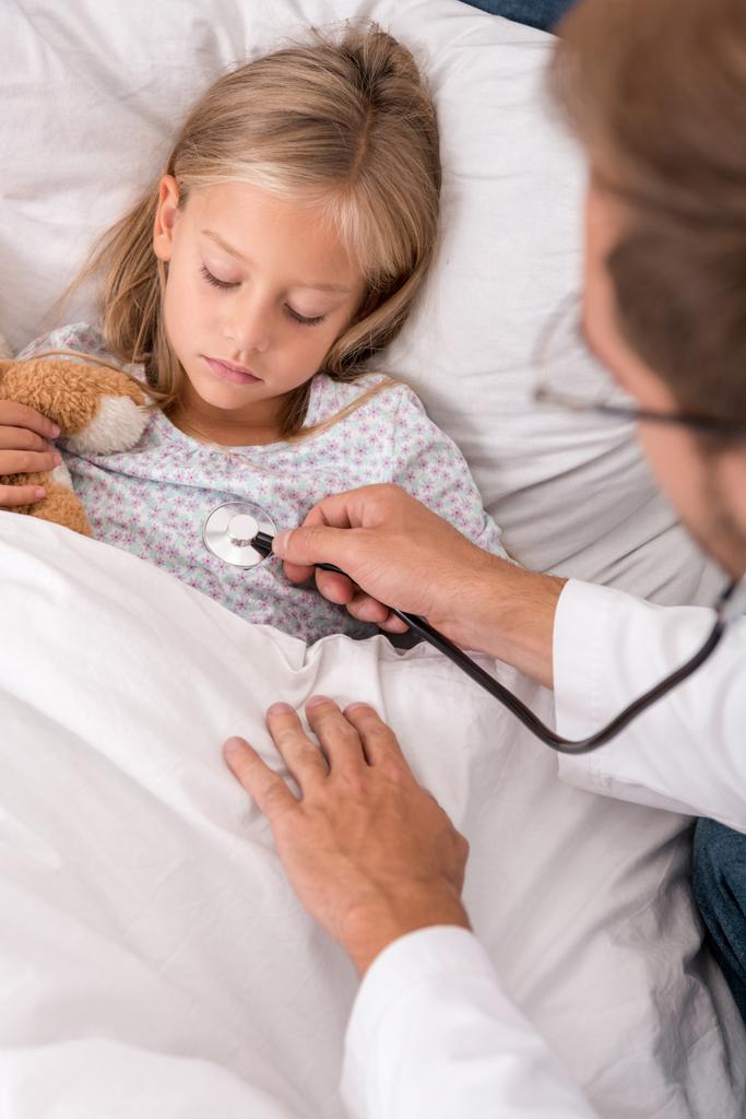 confident pediatrician litening childs breath with stethoscope while she lying in bed - Photo, Image