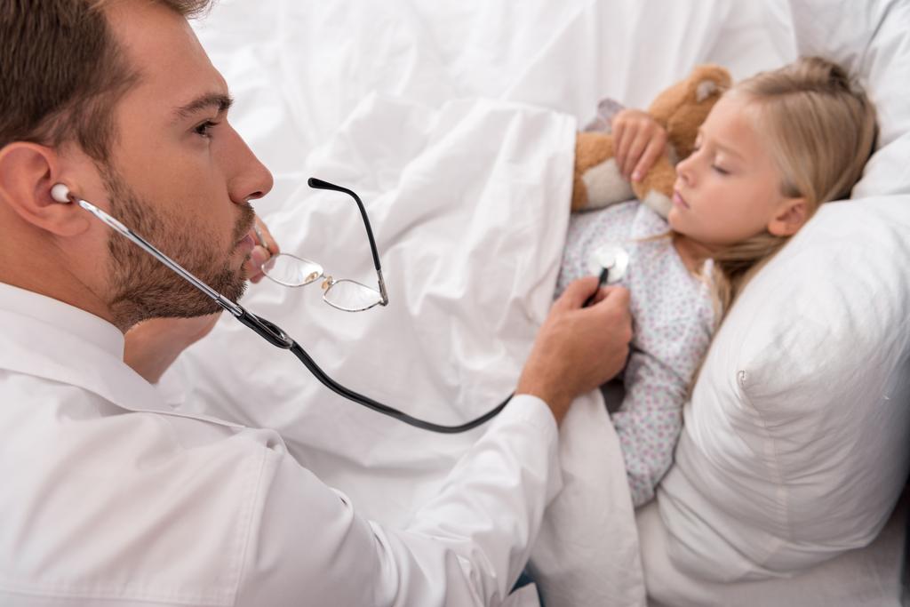 high angle view of confident pediatrician litening childs breath with stethoscope while she lying in bed - Photo, Image