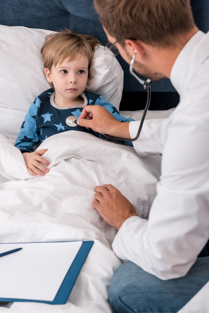 vista ad alto angolo del pediatra litening bambini respiro con stetoscopio mentre si trova a letto
 - Foto, immagini