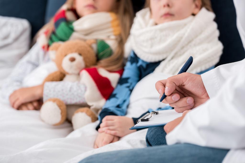 recortado disparo de pediatra escribir en portapapeles mientras está sentado cerca de los niños acostados en la cama
 - Foto, Imagen