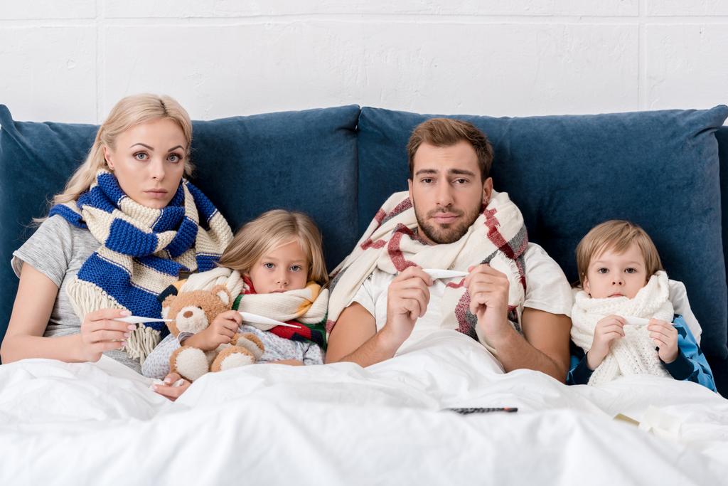 malato giovane famiglia in sciarpe in possesso di termometri elettrici e guardando la fotocamera mentre si trova a letto
 - Foto, immagini