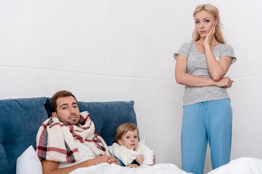 confused woman standing near sick husband and son and looking at camera while they lying in bed - Photo, Image