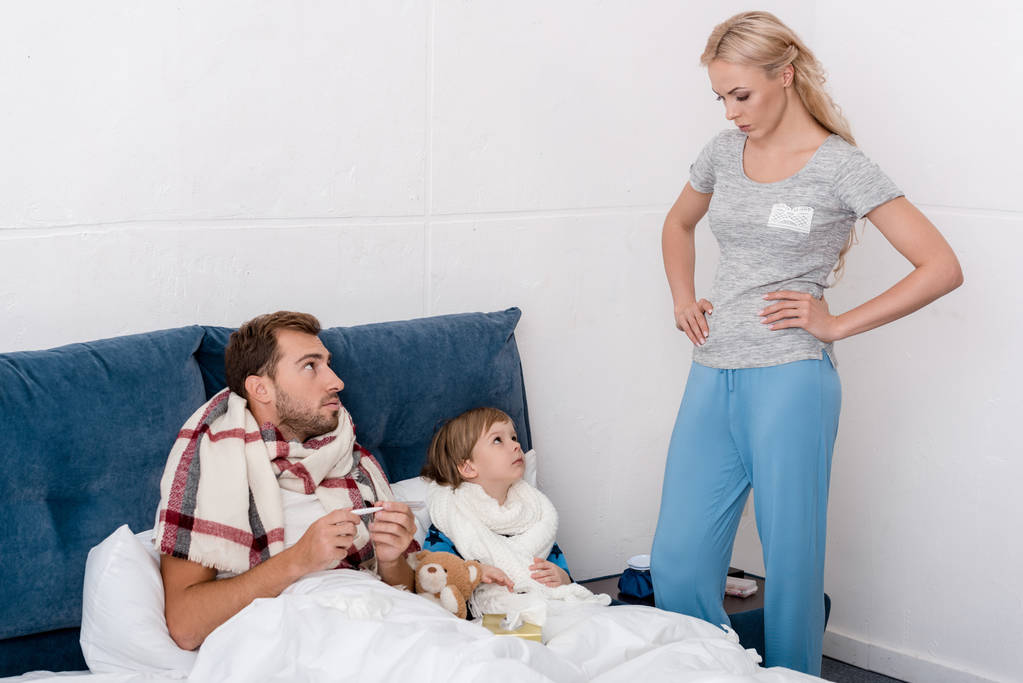 woman standing near sick husband and son while they lying in bed - Photo, Image