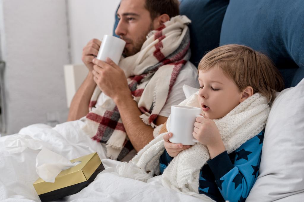 zieke vader en zoon in sjaals met kopjes warm drankje zitten in bed - Foto, afbeelding